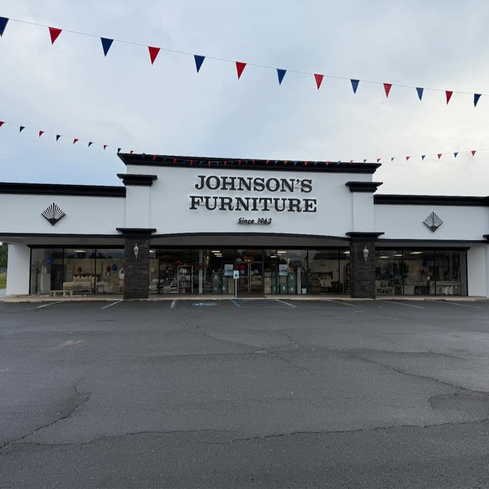 Front exterior of Johnson's Furniture store with a large white facade, dark trim, glass storefront, and red, white, and blue pennant banners strung across the sky above an empty parking lot.